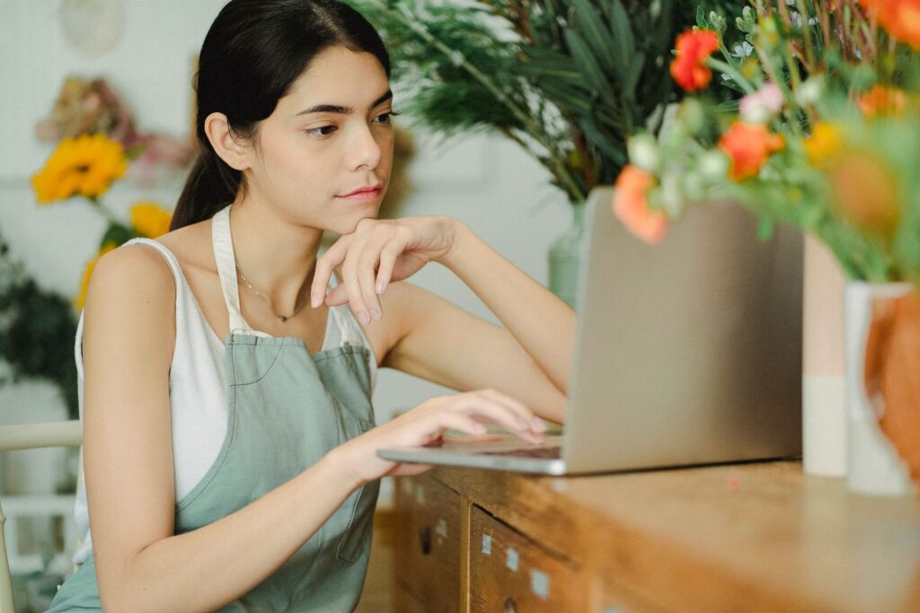 A florist in an apron working on a laptop among flowers in her shop