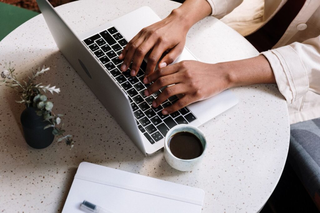 Hands typing on a laptop at a cafe table, with a coffee cup and a notebook