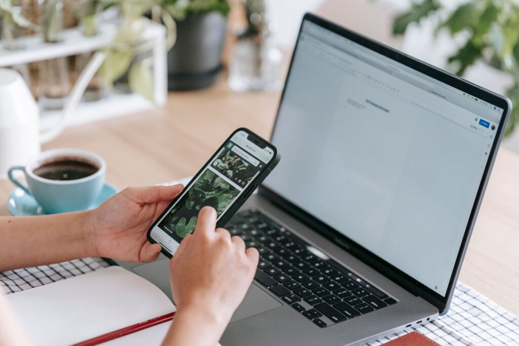 Hands holding a smartphone in front of an open laptop and a cup of coffee