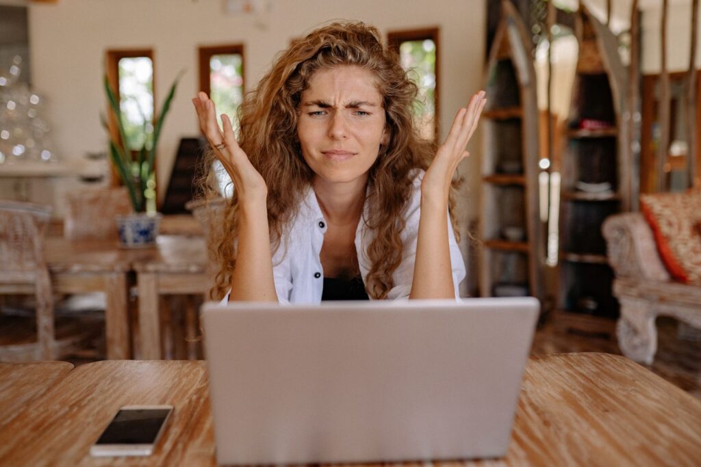 A woman shrugging in confusion at her laptop on a wooden table
